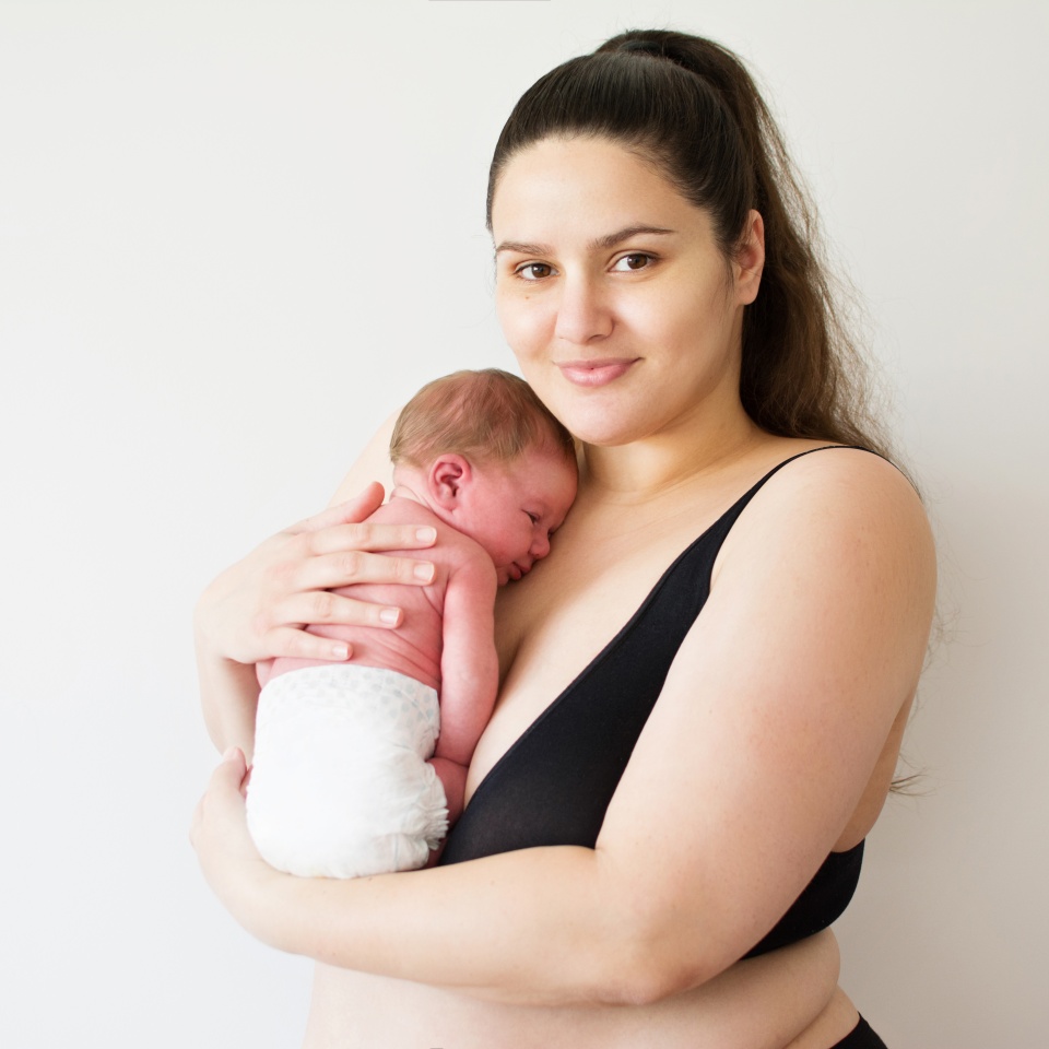 Plus size Caucasian brunette mother smiling and holding her newborn baby. Closeup studio lighting, portrait of mom in black bra and cute baby in a diaper