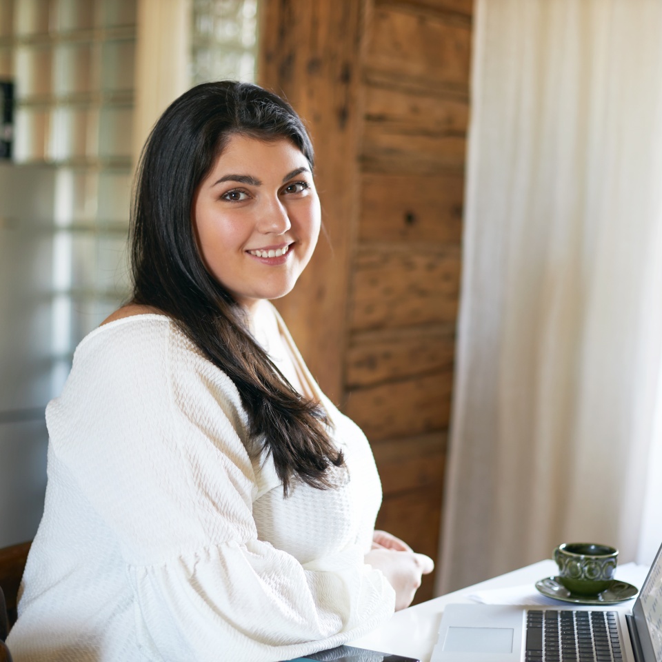Side view of confident skillful young overweight female copywriter or translator sitting at table in front of open laptop, using wireless internet connection for distant work, smiling, drinking coffee