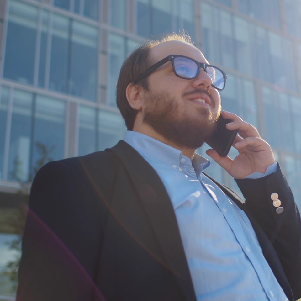 Low angle view of young obese businessman talking on smartphone in city outdoors. Chubby positive employee in glasses having phone conversation standing over business center background