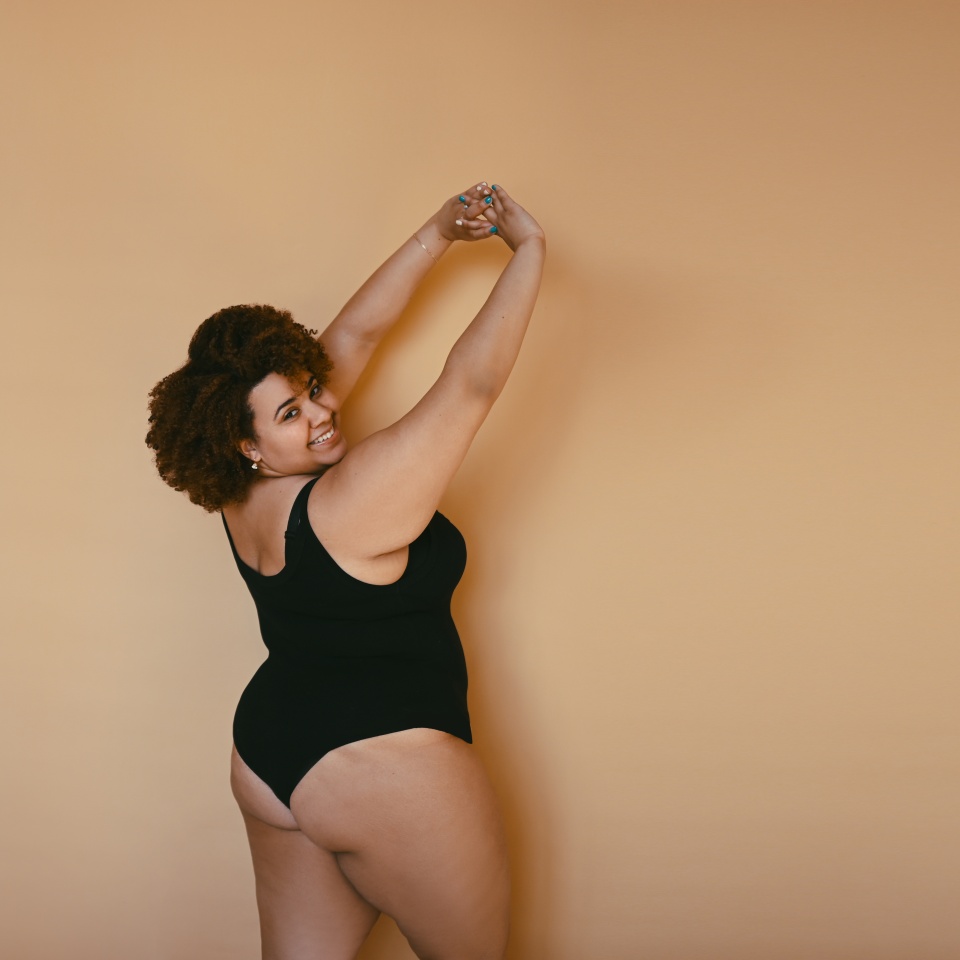 Beautiful curvy oversize African black woman afro hair posing in black bodysuit on beige brown background isolated, body imperfection, body acceptance, body positive and diversity concept. Copyspace