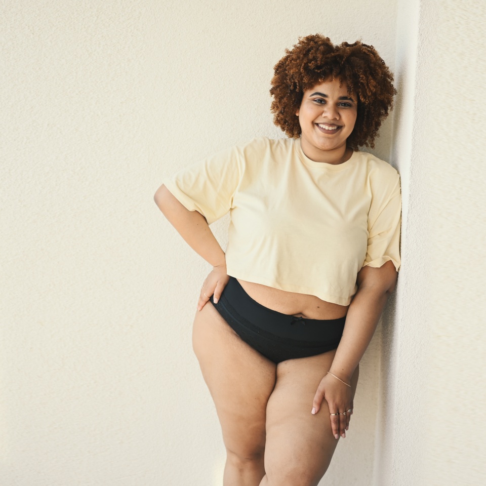 Beautiful happy curvy plus size African black woman afro hair posing in beige t-shirt and black underwear on sunny balcony. Body imperfection, body acceptance, body positive and diversity concept.