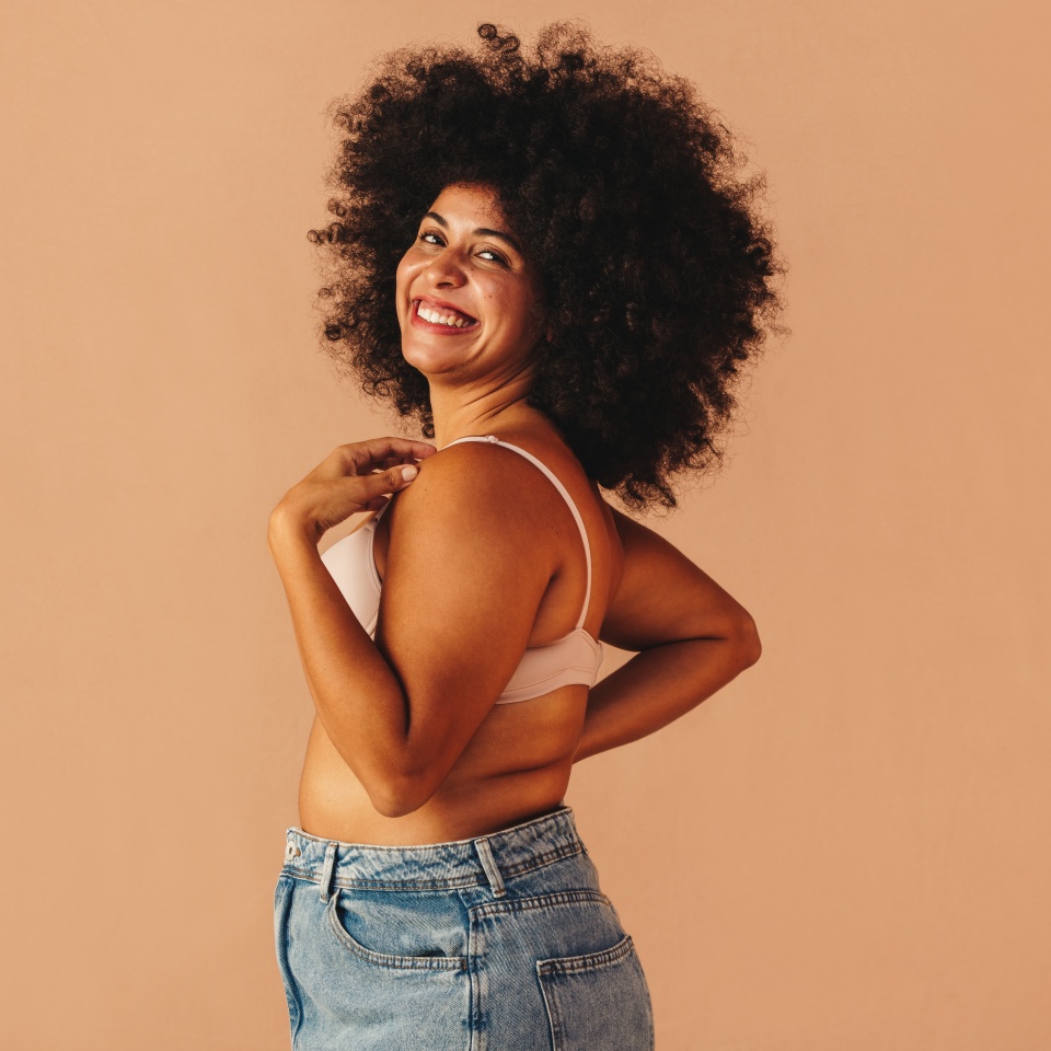 Body confident plus size woman smiling happily while wearing a bra and jeans in a studio. Beautiful young woman with Afro hair embracing her natural body and curves.