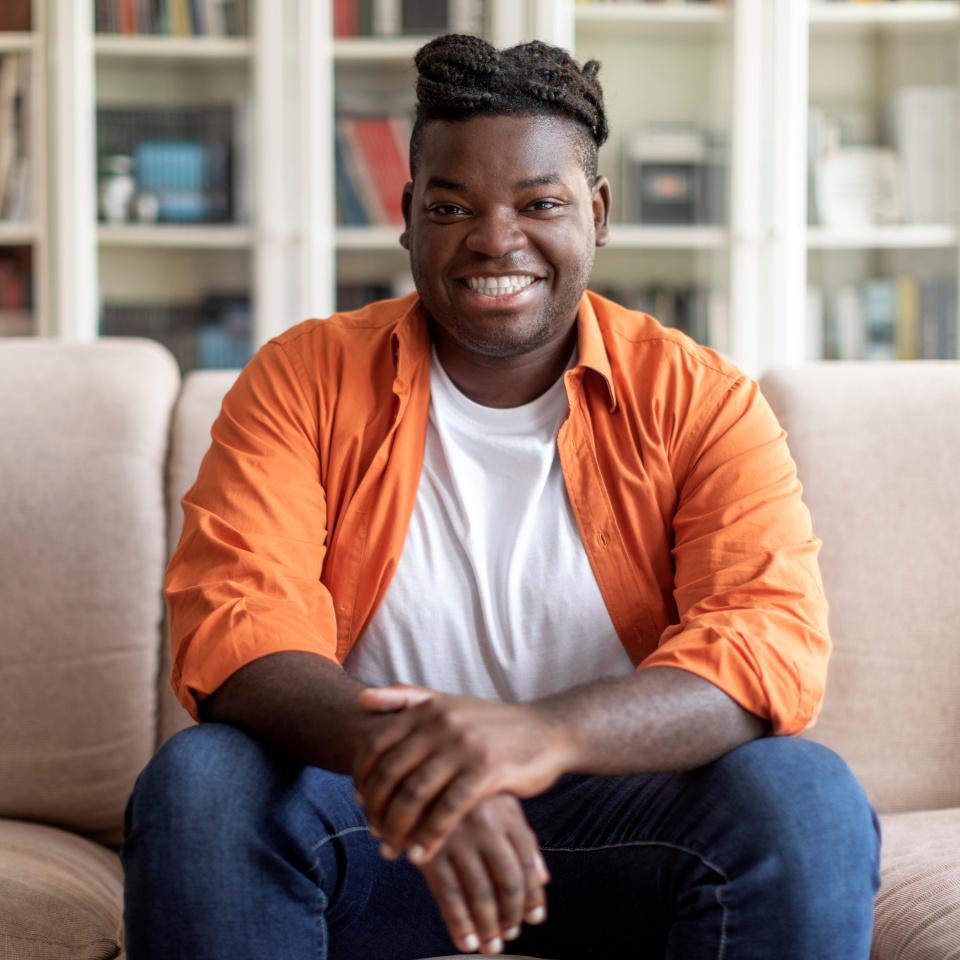Portrait of happy handsome young black man in casual outfit with braids sitting on couch at home, smiling at camera, living room interior, copy space. Millennials lifestyles concept
