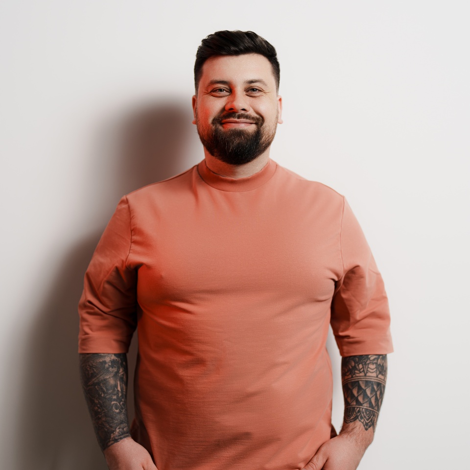 Cheerful young bearded plus sized male model in orange t shirt with tattooed arms looking at camera and smiling against white background in studio