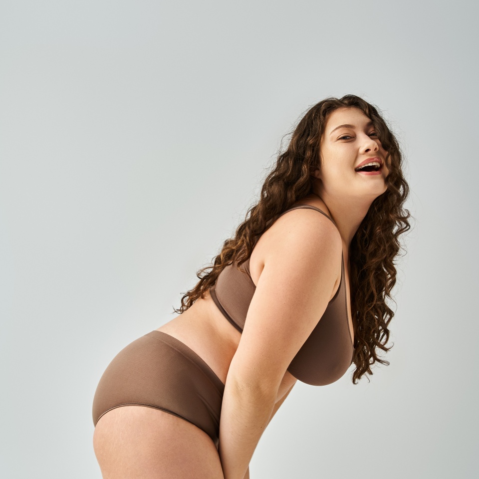 cheerful plus size woman in underwear with curly brown hair laughing and leaning forward sideways