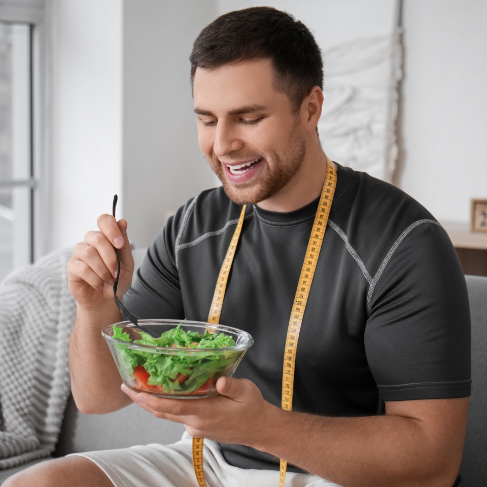 Sporty young man with tape measure and salad at home. Weight loss concept
