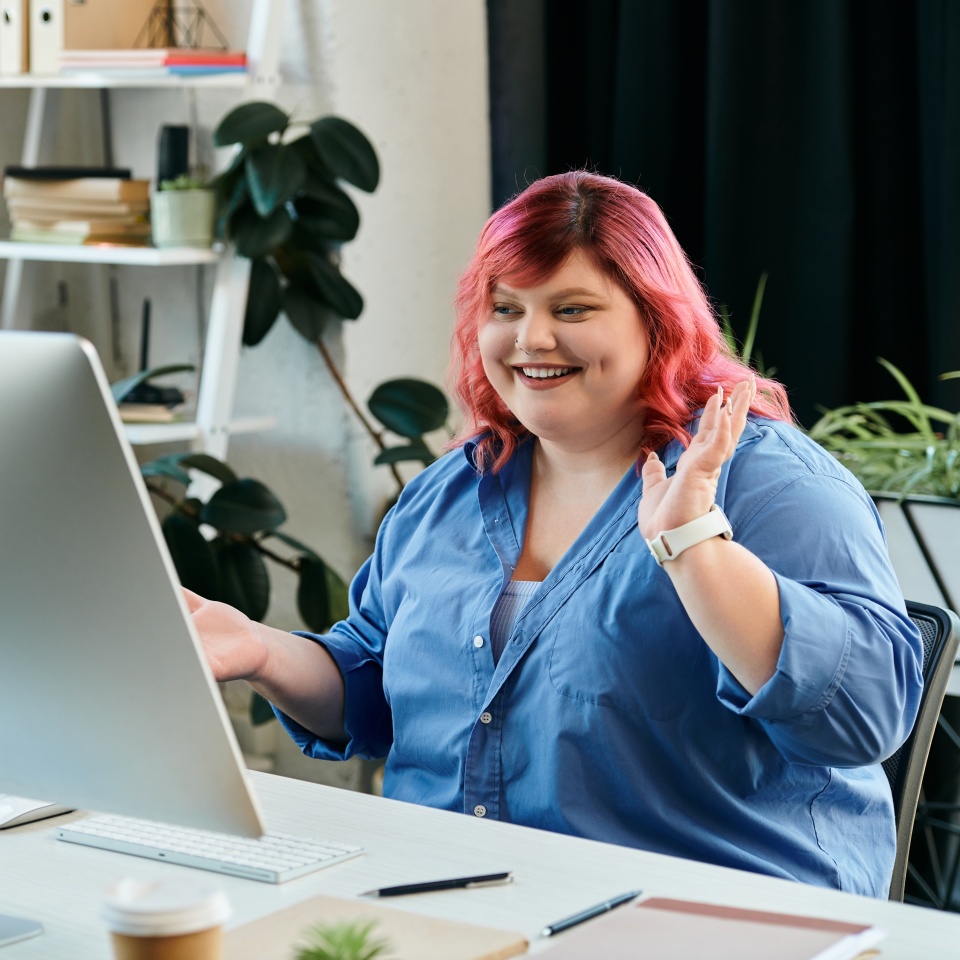 A plus size woman with pink hair waves at the computer screen.