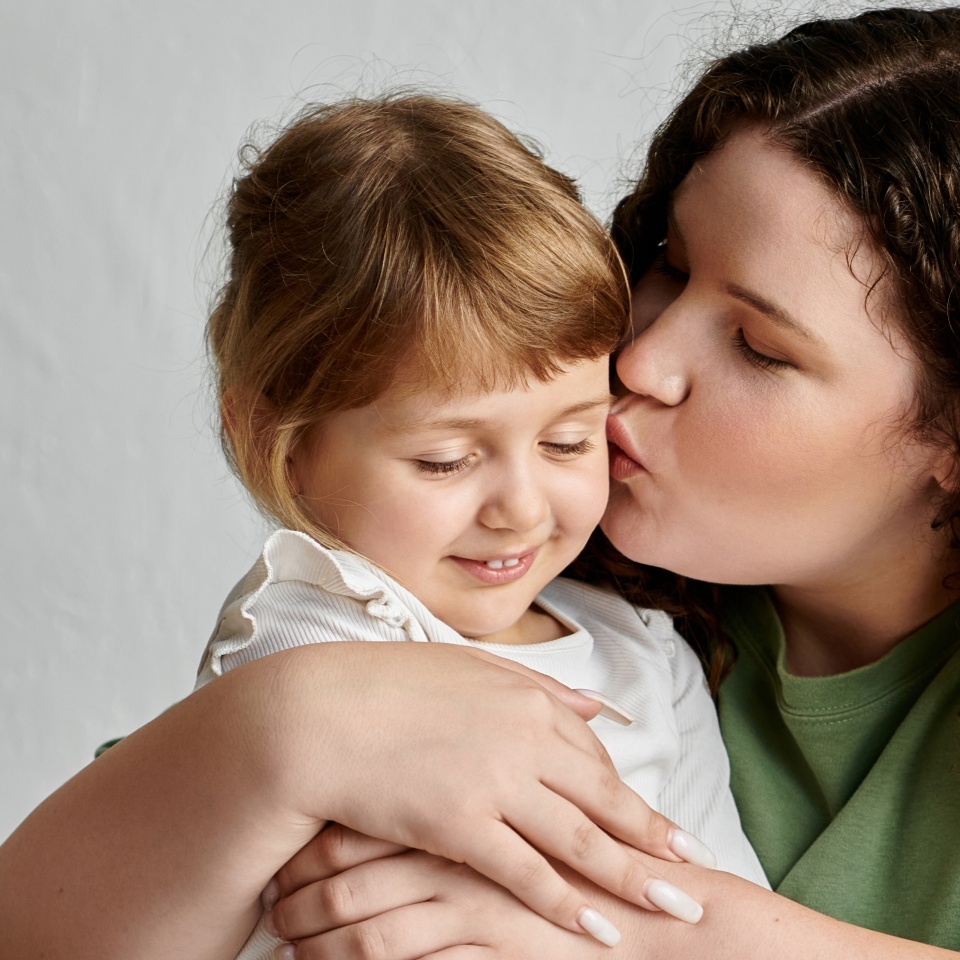 A plus size mother lovingly kisses her daughter, both radiating warmth and happiness together.