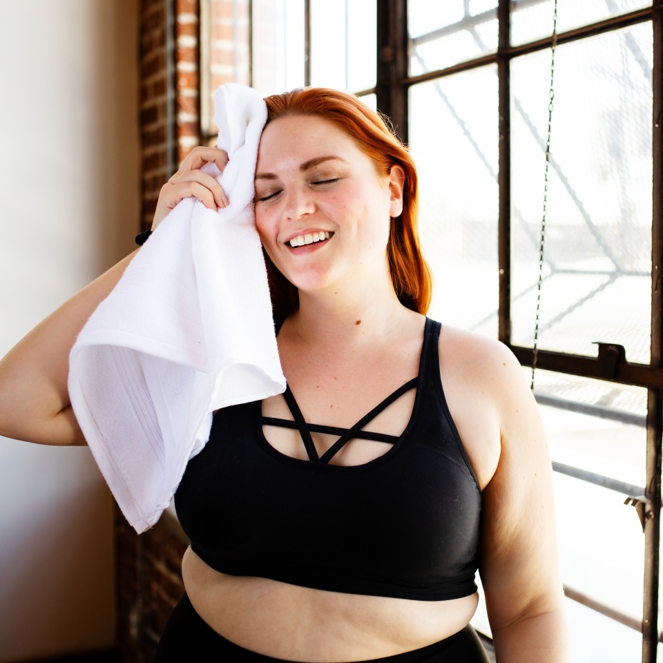 Happy curvy woman with red hair, plus-size, wiping sweat with a towel. Plus-size woman in workout attire, smiling. Red-haired woman, plus-size, cooling down after exercise. Wellness routine.