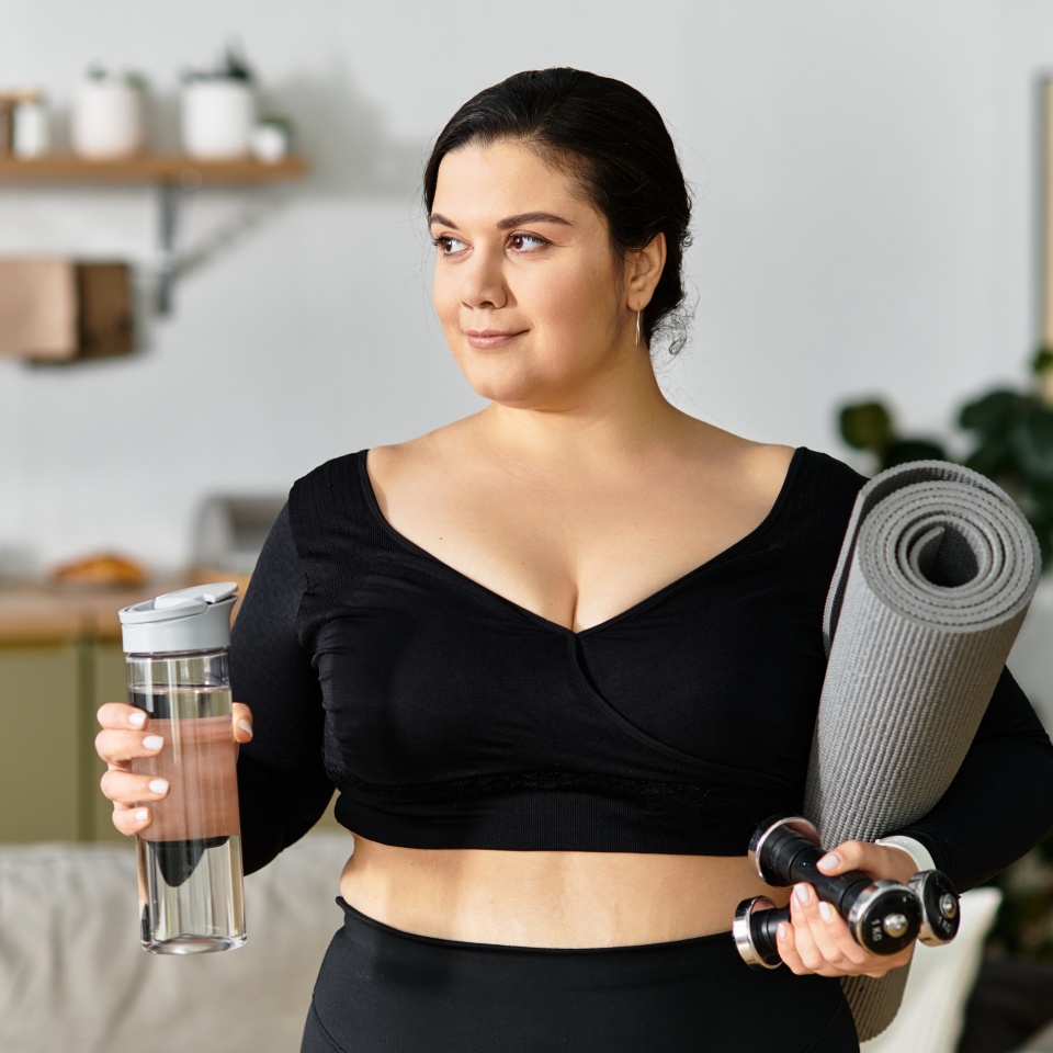 A confident plus sized woman holds weights and a water bottle in a bright room, ready to workout.