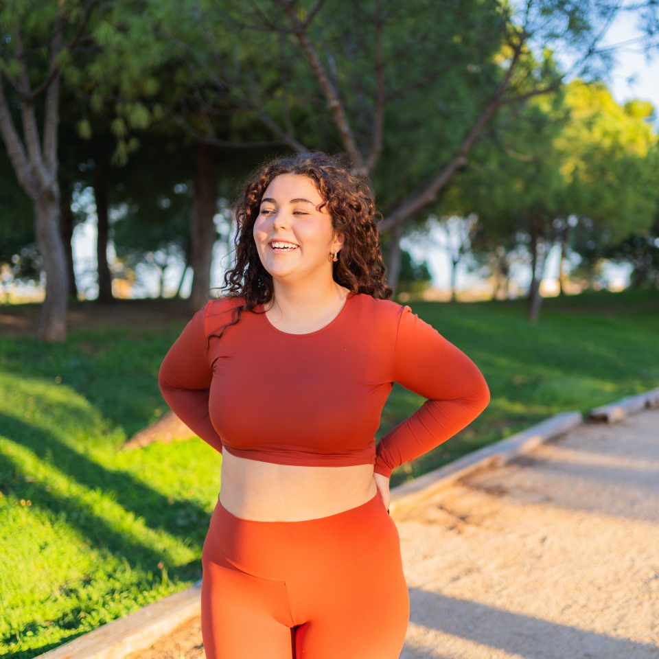 Happy plus size sportswoman wearing orange sportswear standing in a park with hands on hips, smiling and enjoying her workout