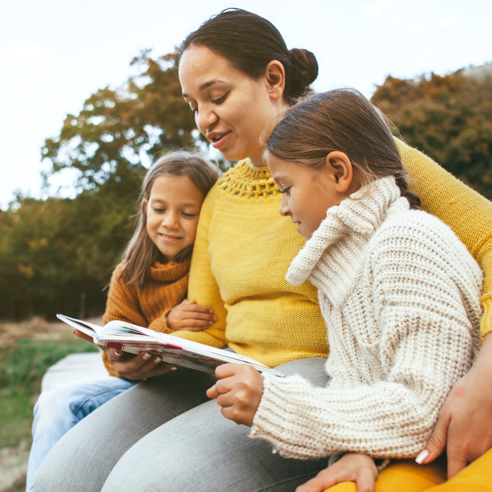 Family spending time together outdoors in autumn. Mom with her daughters reading book in park. Fall weekend in the open air.