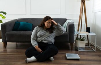 Upset overweight woman sitting on the floor next to scales not understanding why there is no progress in weight loss