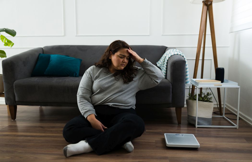 Upset overweight woman sitting on the floor next to scales not understanding why there is no progress in weight loss